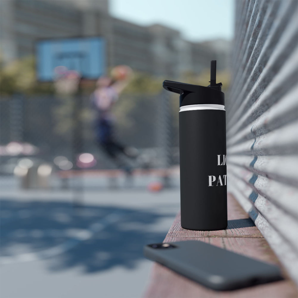 PBP Dad's Survival Kit 18oz. Black insulated water bottle with straw lid on a ledge with a blurred basketball game background